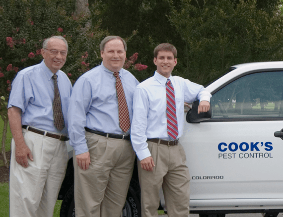 Three smiling men in business casual attire standing next to a Cook's Pest Control company vehicle in a landscaped area.