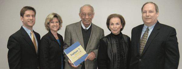 Five Cook's Pest Control founders posing for a group photo indoors; four are smiling and one is holding a document or certificate, all dressed in business attire.