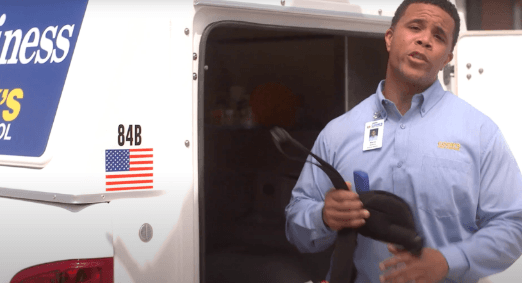 A man in a light blue shirt with a Cook's Pest Control badge is speaking and gesturing, with a van displaying the company's logo and an American flag decal in the background.