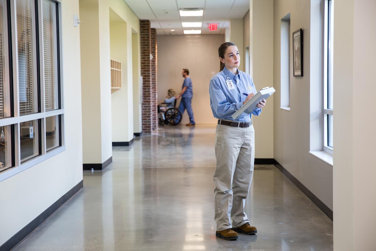 A Cook's Pest Control employee with clipboard in a hallway, with a person in a wheelchair in the background.