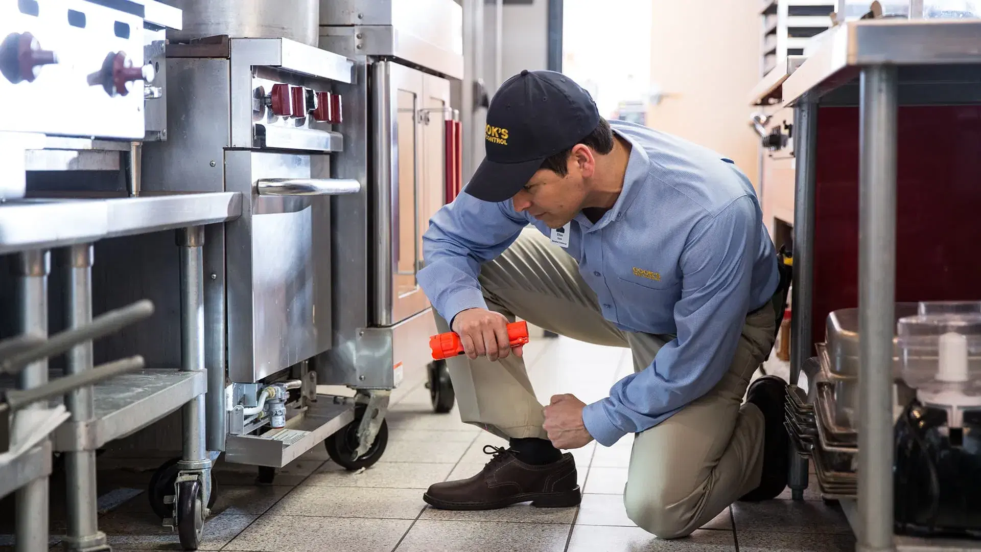 Cook's Pest control technician inspecting beneath commercial kitchen equipment.