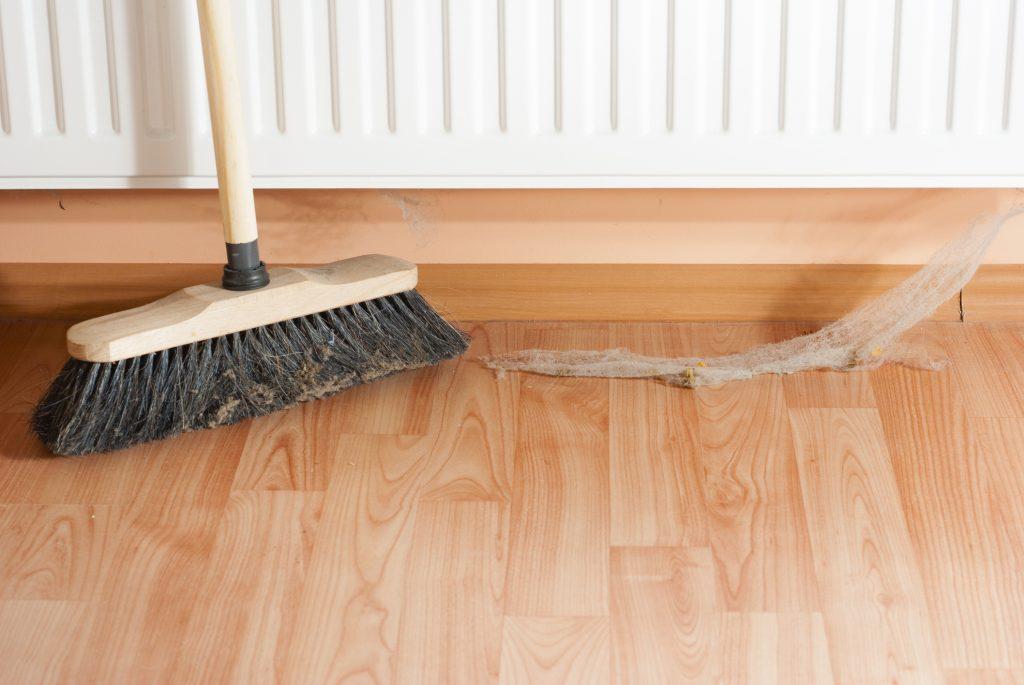 Broom sweeping cobwebs off a wooden floor near a radiator.